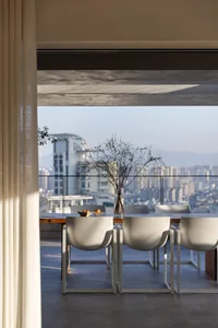 A minimalist living room featuring a concrete floor, white chairs, and a vase with branches, with a cityscape view in the bac