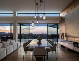 Modern dining room with glass table, upholstered chairs, and wooden floors, featuring large windows overlooking a sunset.