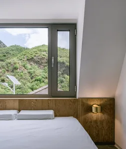A minimalist bedroom featuring a bed with white sheets and a wooden headboard, overlooking a green hillside through a large w