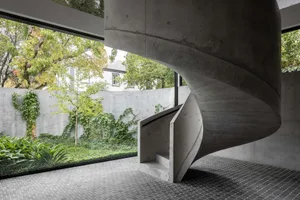 A modern, industrial-style spiral staircase made of concrete with a glass railing, surrounded by greenery and large windows.