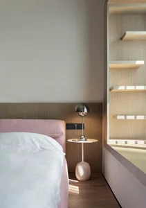 A minimalist bedroom with a pink headboard bed, white bedding, and a wooden nightstand with a round table lamp.