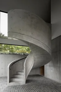 Circular concrete staircase with a dark rail in an industrial space with large windows.