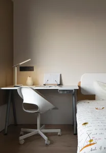 A modern minimalist bedroom with a white desk and chair, a bed with a white and floral comforter, and a white desk lamp.