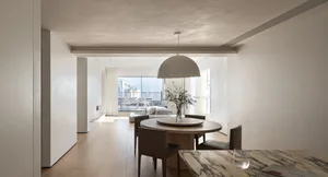 A minimalist dining room with a round wooden table and chairs, accented by a modern hanging light fixture and a vase with flo