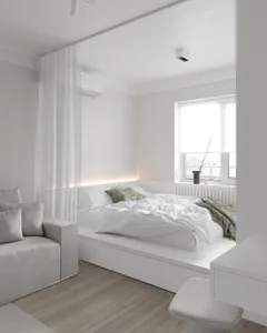 Modern, minimalist bedroom with white walls, furniture, and bedding, featuring a window, a small sitting area, and a low-prof