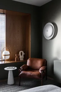 A modern living room featuring a brown leather armchair, a white textured side table, and abstract sculptures on a wooden win