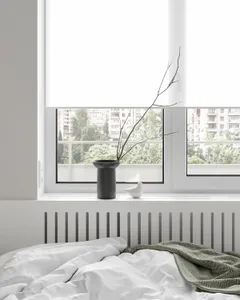 A minimalist bedroom with a white blanket and gray blanket on a bed, featuring a vase with a branch and a bird figurine on a 