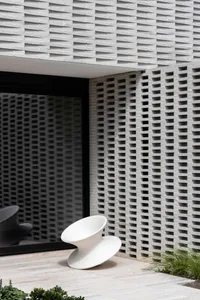 A modern exterior design featuring a concrete wall with a grid pattern and a unique, angular white chair in front of a large 