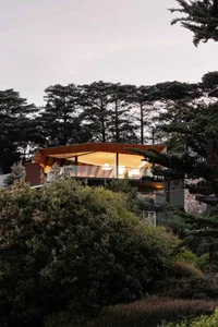 A modern house with a glass wall and wooden frame surrounded by greenery.