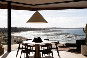 A modern residential dining room featuring a wooden table and chairs with a view of the beach and ocean.