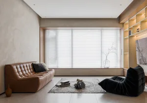 A modern minimalist living room with a leather sofa, a black upholstered chair, a rug, and a window with blinds.