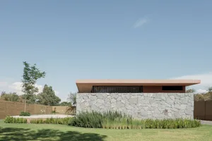 Modern house with stone and concrete exterior, surrounded by greenery and a clear blue sky.