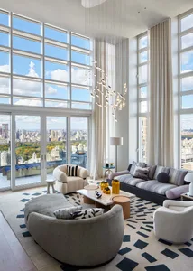 Modern living room with large windows overlooking a cityscape, featuring contemporary furniture and a chandelier.