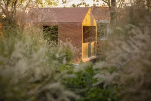 A minimalist brick house with a small window and a simple roofline surrounded by tall grasses and trees.