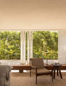 A minimalist living room with natural light, featuring a wooden chair and side table against a wall of windows.