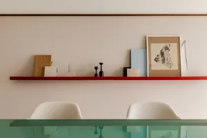 A minimalist home office with a red shelf displaying various objects, including a framed abstract piece, small sculptures, an