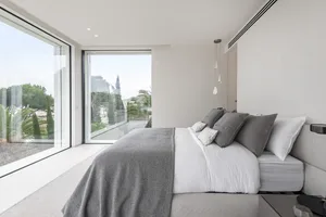 A modern, brightly lit bedroom with a bed dressed in light fabrics and glass windows.