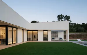 Modern residential backyard featuring a white concrete structure with glass sliding doors and a green lawn.