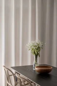 Modern dining room featuring a wooden table and a vase of white flowers on a glass table, set against a beige curtain backgro
