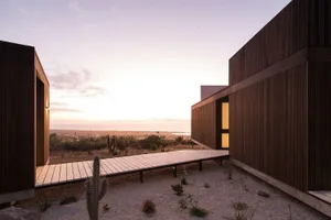 A minimalist modern house with wooden exterior walls and a sandy ground, showcasing the exterior design and social space.