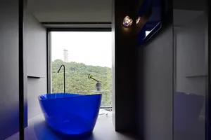 A minimalist bathroom with a bright blue glass tub, a vase with a flower, and a window overlooking a lush green hillside.