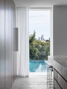 A modern residential kitchen features a glass window overlooking an infinity pool and city skyline, with marble countertops a