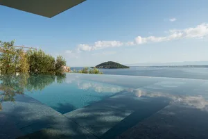 A minimalist swimming pool with a concrete deck and glass walls overlooks a calm sea.