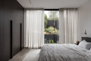 A minimalist bedroom featuring white curtains, a wooden floor, and a white bed with a view of greenery outside.