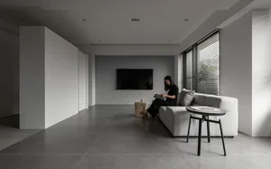 A modern minimalist living room with concrete flooring, a white sofa, and a reading woman sitting beside a window with blinds