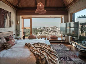 A modern minimalist bedroom with a large window showcasing a town view, a bed with a fur blanket and colorful pillows, and a