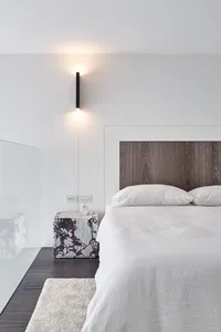 Modern minimalist bedroom featuring a wooden headboard against a white wall, illuminated by a wall-mounted sconce, with a whi