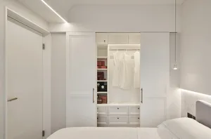 A minimalist bedroom with white walls, a bed, and a built-in closet with white doors and white hanging garments.