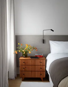 A modern bedroom featuring a wooden nightstand with a vase of flowers, books, and a small lamp.