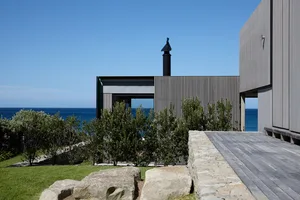 Modern beach house with wooden slat exterior and concrete base, featuring a chimney and glass windows.