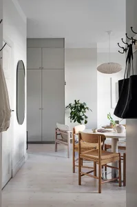 A minimalist dining room with wooden chairs and a white table, featuring a potted green plant and a round pendant light.