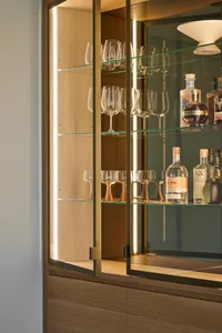 A modern bar cabinet featuring glass shelves and LED lighting, displaying a variety of wine glasses and bottles.