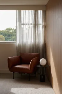 A brown leather armchair next to a window with sheer curtains, featuring a spherical lamp on a side table.