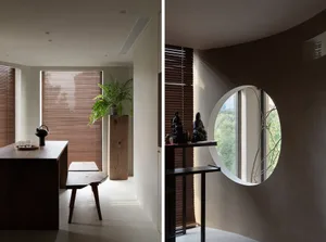A minimalist living space features a wooden table, bench, and plant under brown blinds, with a circular window and additional