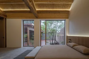 A minimalist bedroom features a timber frame ceiling and large windows overlooking a garden.