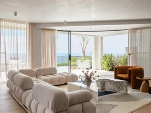 A modern minimalist living room with a large tufted sofa, marble tables, and floor-to-ceiling windows overlooking the ocean.