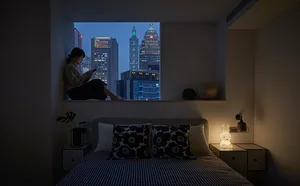 A modern bedroom with textile bedding and a glass window overlooking a city skyline.