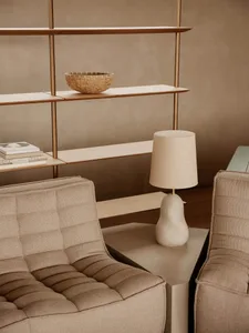 A minimalist living room featuring beige couches and a table lamp with a beige shade, with a beige bookshelf in the backgroun