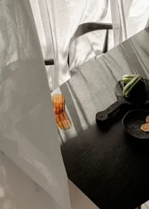 極簡室內設計，黑色餐桌搭配玻璃花瓶與一碗綠色蔬菜，置於薄紗窗簾前