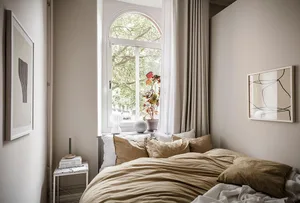 A cozy minimalist bedroom featuring a bed dressed in beige textiles, a small side table, and a window with white curtains ove