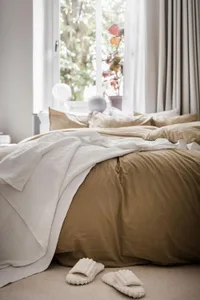 A minimalist bedroom with a bed covered in tan-colored cotton towels. White slippers are placed on the floor.