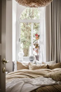 A minimalist bedroom featuring a bed with beige bedding, a potted plant, and sheer curtains.