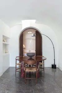 A minimalist dining room with a round table made of red marble and wooden chairs surrounded by white walls and dark grey marb