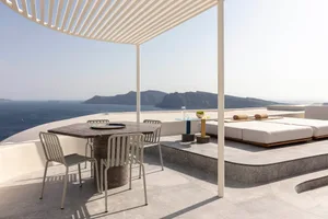 A minimalist residential patio featuring a cement table, metal chairs, glass plant holder, and metal pergola with a view of t