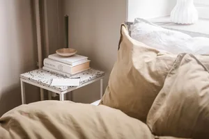 A modern bedroom featuring white textured cotton sheets on a bed, a small side table with books and a candle holder, and a wi
