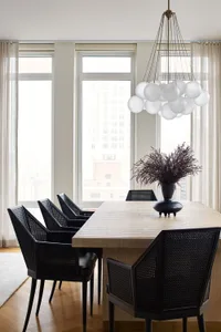 A modern dining room featuring black metal chairs around a wooden table, with a chandelier and floor-to-ceiling windows.